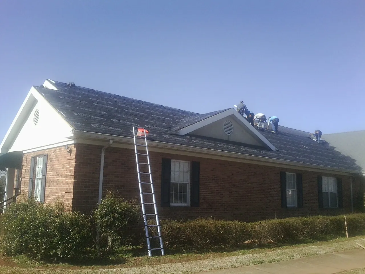 Crew tearing shingles off a brick ranch roof with a ladder set — Emergency Roof Repair work by our crew in Colona