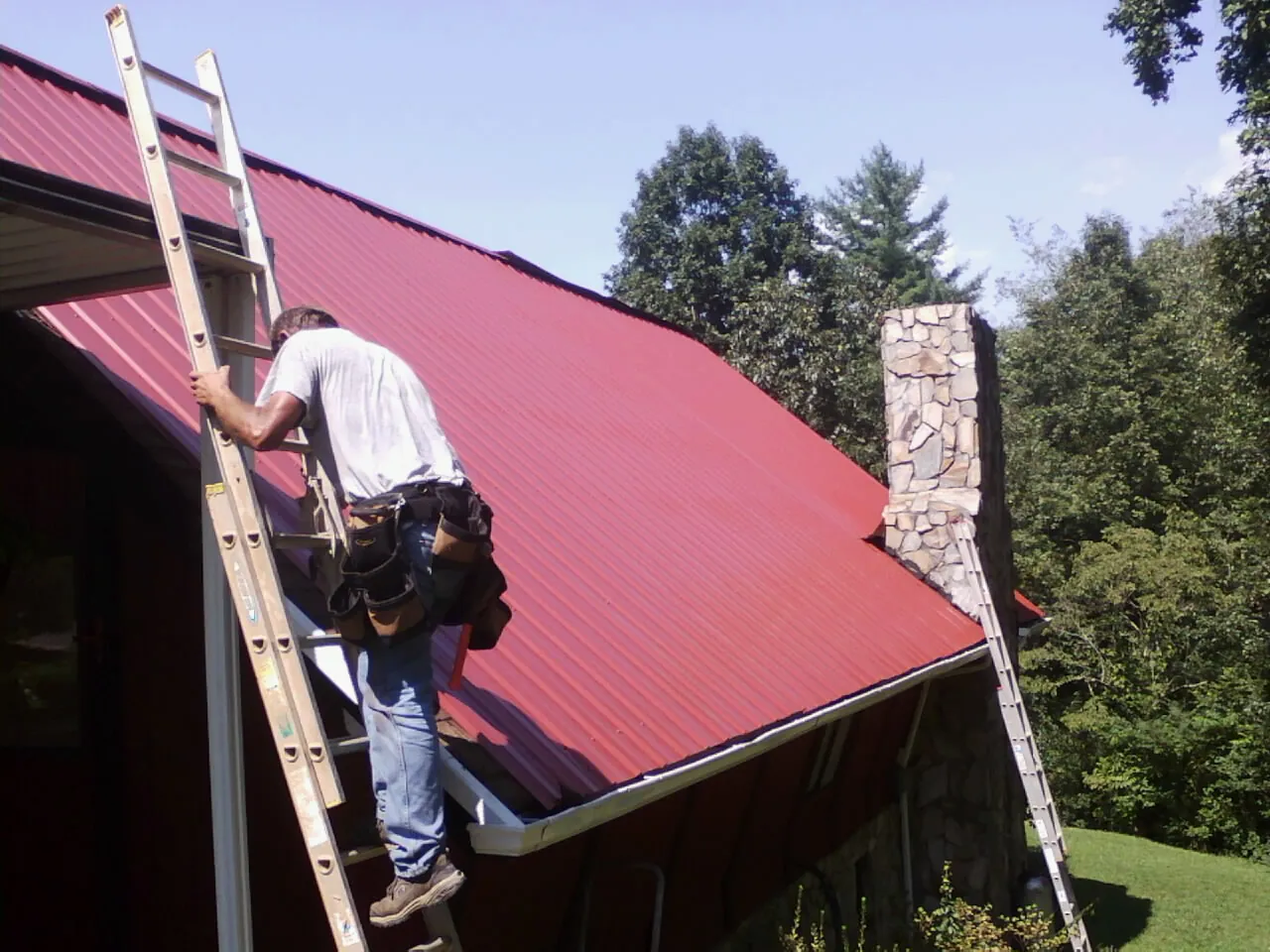 Roofer on a ladder beside a red metal roof and stone chimney — Soffit & Fascia Repair by Roofing Brothers in Colona